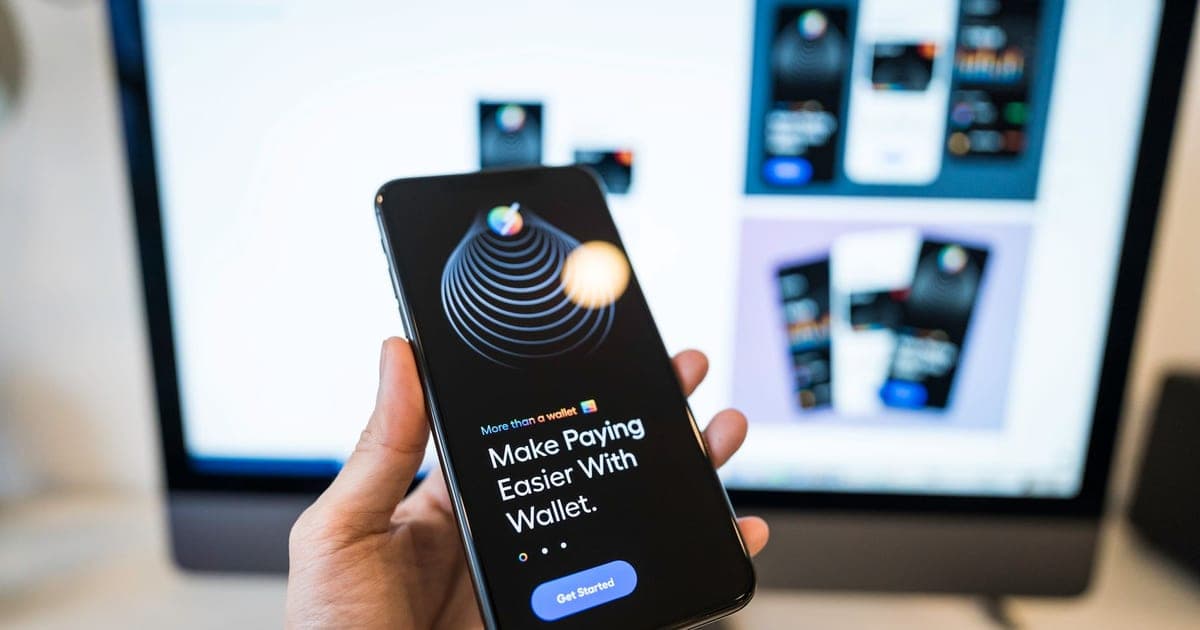 Hand holding smartphone displaying digital wallet app interface, blurred monitor in background.